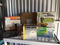 Multiple houseware items displayed on metal rack including boxed iced tea pot, Black & Decker kettle, Hamilton Beach SaladXpress, Swiffer cloths, transparent pitcher, black box