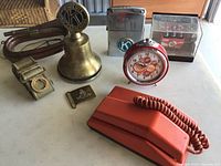 Overview of the lot items including vintage telephone, red alarm clock, brass bell, lighter, belt buckles, belt, and glass display with model vehicles.