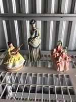 Three porcelain figurines placed on metal surface: two identical Lefton China Bo Peep seated girls holding staffs, and one Royal Doulton standing woman figurine in green and cream dress.