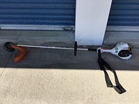 Full side view of trimmer showing engine housing, shaft, handle, strap, guard and head laid on concrete