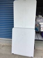 Two Lifetime white folding tables leaning against a storage unit, showing rectangular shapes and edges with visible use marks.