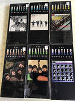 Overview of all 6 Beatles compact discs with original cover art, laid flat in two rows. Disc cases show mostly black background with album covers and 'The Beatles Compact Disc' text. Most are still wrapped in plastic with condition appearing used and new.