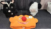 Three mascot heads displayed together on a dark surface, showing a black and white skunk, white and black panda head, and an orange Ernie mascot head with a large red nose.