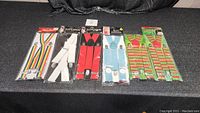 Full view of all 6 pairs of suspenders laid out horizontally, still in plastic packaging, showing different colors and patterns.