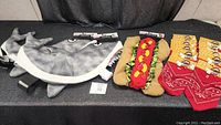 Photo showing two grey shark dog costumes with fins and teeth detail, one brown hot dog costume with lettuce and mustard detail, and four red cowboy dog bandanas in packaging.