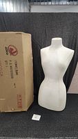 White female torso mannequin standing next to original box on black backdrop.