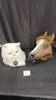 Both masks placed on a black textured surface with a dark backdrop, showing the horse and white cat masks side by side.