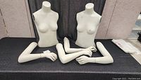 Two female mannequin torsos and six mannequin arms arranged on black cloth surface.