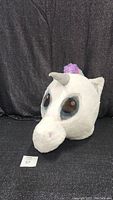 Front view of the white unicorn mascot head with large brown eyes and gray horn, purple and blue mane visible.