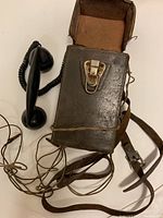Full view of the antique military field telephone with leather carrying case, coiled wires, and handset positioned to the side.