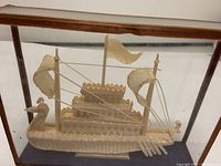 Front view of the Asian dragon boat carving showing the dragon head prow, central flags, and oars in a glass display case with wooden frame.
