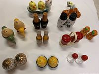 Photo of eleven pairs of vintage salt and pepper shakers arranged on a white surface showing diverse shapes and materials such as plastic, ceramic, glass, wood, and tin. Various quirky designs including birds, chickens, corn, and zodiac motifs.