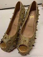 Pair of gold high heel pumps with peep toe and gold spikes across upper surface, showing sequins and embellishments.