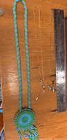 Handmade beaded necklace in blue and green with circular pendant and fringe lying flat next to two thin gold tone necklaces with small pendants, measured by ruler.