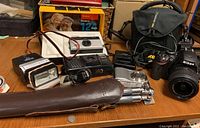 Overview of multiple cameras, Nikon D70 DSLR with attached lens, tripod in leather case, multiple flashes and accessories on wooden table.