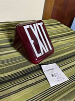 Red glass exit lamp shade sits on green and black striped fabric, showing front side with white EXIT text and a 7-inch tall and 3.25-inch diameter tag nearby.