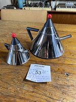Two conical stainless steel tea pots with black angular handles and red spherical knobs on lids placed on wooden table.