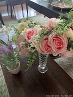 Overall view of the four artificial flower arrangements displayed together on a wooden table with chairs and carpet background. The largest arrangement shows pink roses with green leaves and white accent flowers in a clear textured vase.