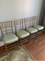 Four gold Chiavari chairs with green seats arranged in a row on wooden floor, showing overall condition and style.