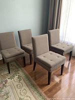Full view of four matching dining chairs arranged on rug near window with beige/taupe button-tufted fabric upholstery and dark wooden legs.