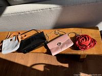 Four small purses displayed in sunlight on wood surface: a gray tote with tan straps, black patent purse, pink purse with metallic clasp, and red rose-shaped purse.