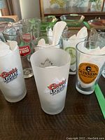 Eight branded beer glasses arranged on wooden table, two Coors Light SubZero frosted glasses, two Guinness Extra Stout clear glasses, plus other brands visible.