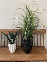 Two artificial plants side-by-side on a wooden chair back: taller grass-like plant in black pot and shorter broad-leaf plant in white pot.