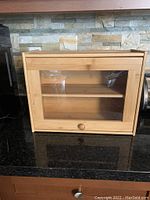 Front view of wooden bread box with glass panel and wooden knob on countertop.