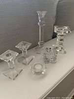 Six crystal candle holders placed on a white surface in front of a gray couch, showing variety in shape and size, emphasizing transparency and light reflection.