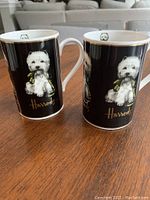 Two white china coffee mugs with black background and Westie dog illustrations sitting on a wooden table.