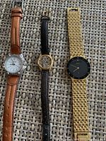 Front view of three ladies wristwatches showing dials and straps: Citizen with white dial and brown leather strap, Swiss with gold-tone oval dial and black leather strap, Seiko with black dial and gold-tone bracelet.