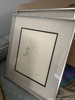 Photo showing two framed prints leaning against a wall, both featuring minimalist line drawings of women with foliage, with light fabric mats and silver frames.