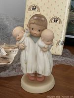 Porcelain figurine of a young girl in white nightgown holding two babies, placed in front of original box and bubble wrap.