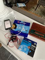 Photo showing iPhone with 'Hola' welcome screen, Verbatim CD-R discs box, Epson ink cartridge, wired earphones, and wooden pen set