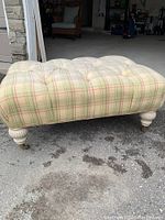 Photo showing plaid upholstered ottoman with tufted cushion and turned wooden legs on pavement.
