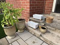 Full lot showing seven pieces of planters arranged on patio steps and ground. Includes two large ceramic pedestal planters with plants, three small round pots, two white rectangular planters, one small metallic pot and one small woven basket.