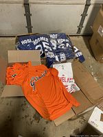 Four open boxes showing orange Giants shirts, white Indians shirts, and blue Hosmer shirts