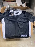 Open box showing one black and white Maverick lacrosse shirt unfolded on top, showcasing the front design and logo tag on collar and bottom hem.