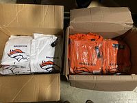 Two open cardboard boxes containing folded men's Denver Broncos Majestic T-shirts in white and orange colors. The shirts bear the Broncos logo and name and have tags attached, indicating they are new.