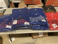 Plastic-packaged long sleeve MLB T-shirts folded on table, showing Mets, Phillies, Royals team logos and tags.