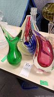 Three art glass vases displayed on a table with a green vase, striped vase with a stopper, and pink-purple vase