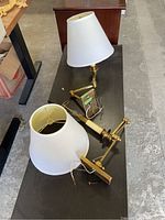 Two brass wall lamps placed on a table, showing full view including white fabric shades, brass articulated arms, and exposed wiring.