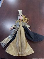 Full view of a doll with gold textured gown, black and gold patterned robe, and tall silver ornate headdress on a dark surface.