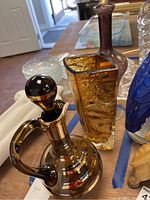 Photo showing an amber glass stoppered jug with handle, a square amber textured vase, and a tall purple glass vase with bulbous body on a clear foot.