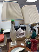 Pair of two decorative table lamps with different pleated shades, one beige with gold trim and one cream with blue trim, textured clear glass with yellow tint bases on stone square bases with gold detail.