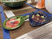 Two Moorcroft ashtrays on a brown quilted cloth. One green teardrop-shaped with pink-red flower, one dark blue triangular with red flower.