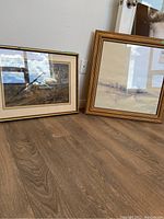 Two framed watercolours leaning against the wall on a wooden floor, one showing a winter landscape, the other an industrial scene with a crane.