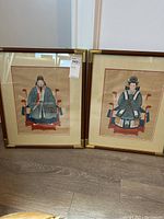 Two framed prints side by side on the floor, showing man and woman in traditional Asian dress, with brown and gold frames.