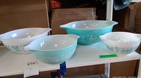 Four Pyrex bowls arranged on a shelf. Bowls have a blue exterior with white colonial pattern and white interior.
