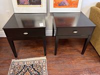 Front angle view of two matching dark wood side tables with drawers and metal pulls, placed against a wall with framed artwork above.